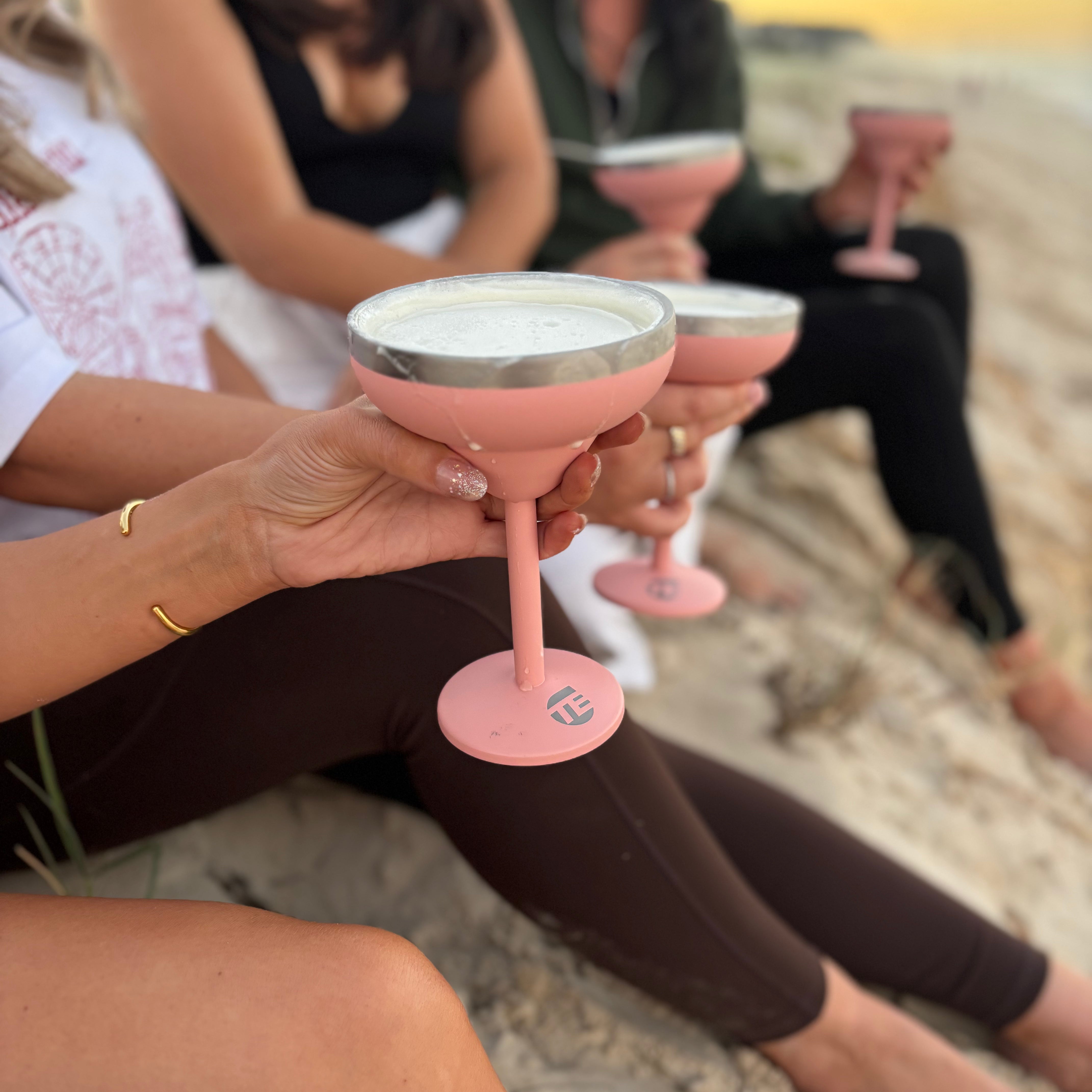 People sitting on a beach holding pink cocktail glasses with a sunset in the background.
