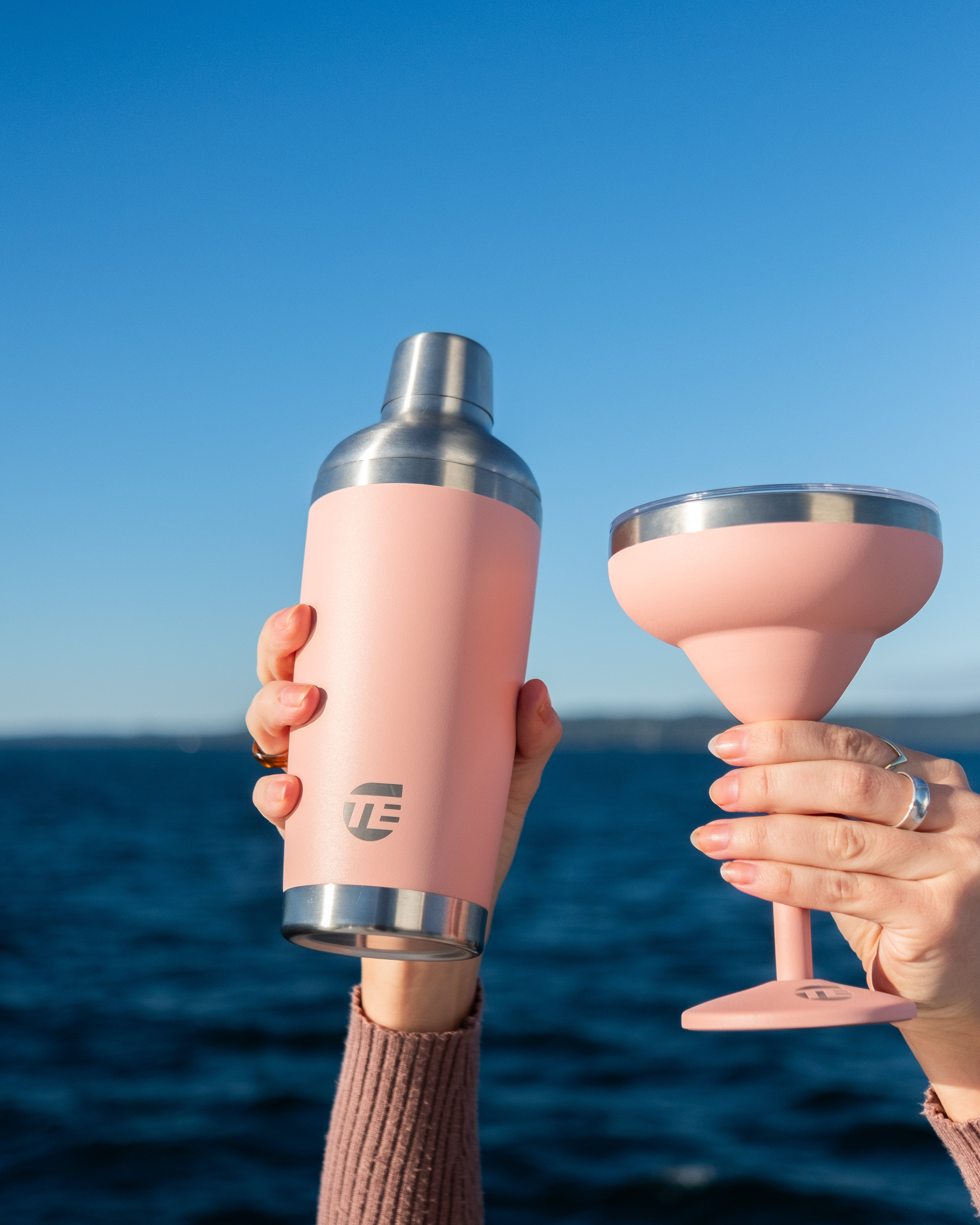 Two pink drink insulated drink ware items held against a blue sky and ocean background