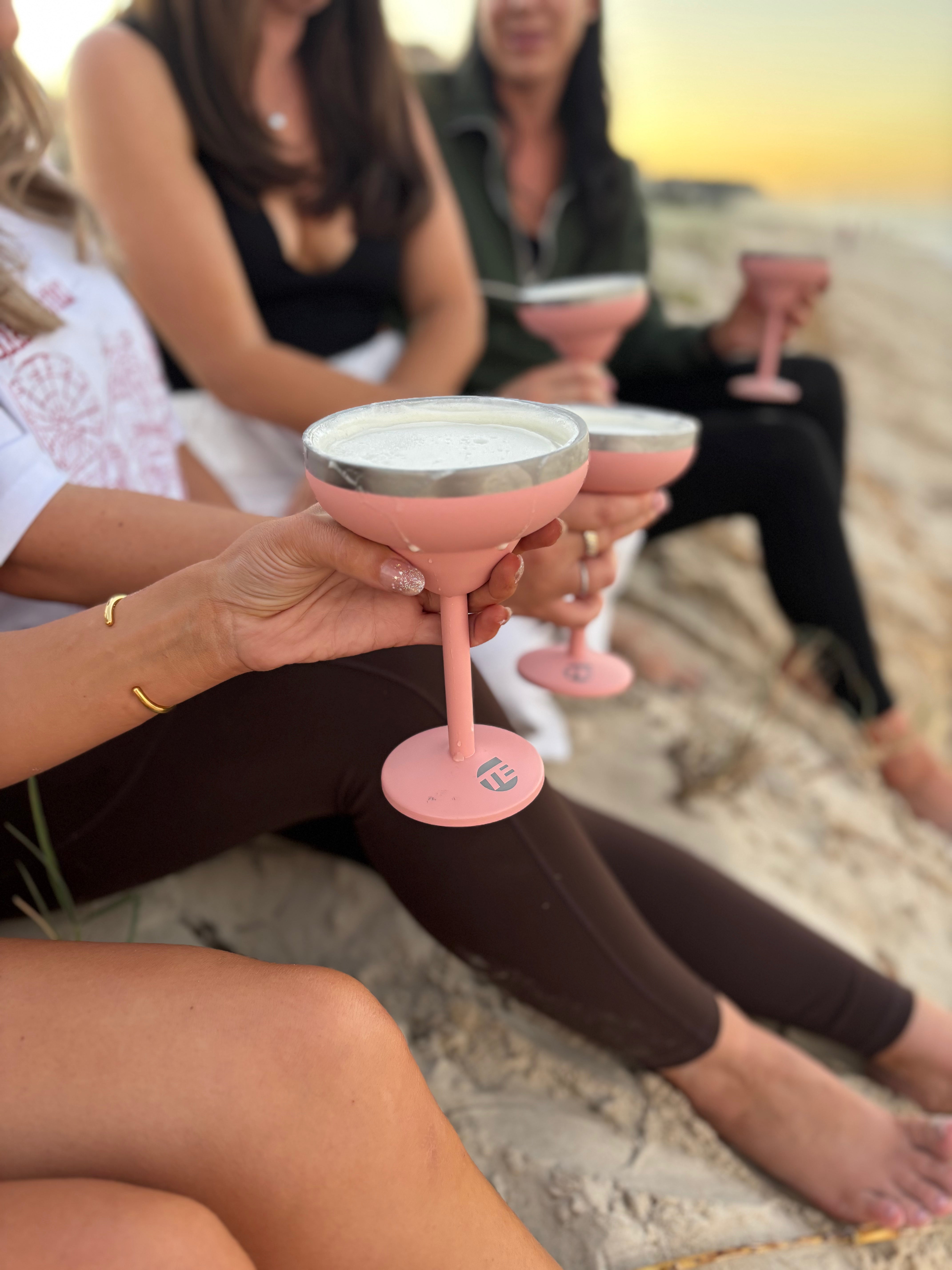 People sitting on a beach holding pink cocktail glasses with a sunset in the background.