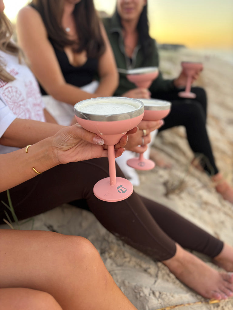 People sitting on a beach holding pink cocktail glasses with a sunset in the background.