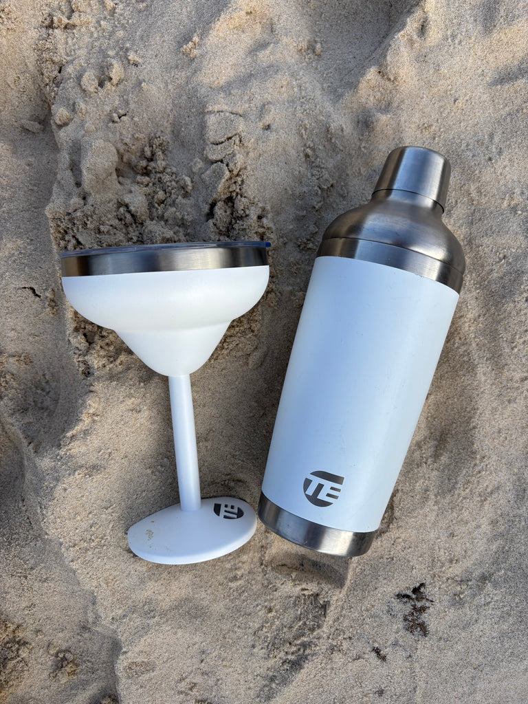 White cocktail shaker and glass on sandy surface